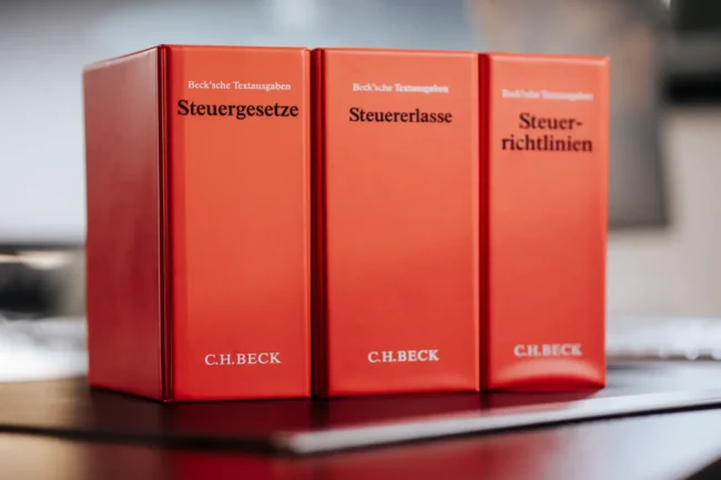 Drei rote Rechtsbücher zum Steuerrecht, Steuererlasse und Steuerrichtlinien stehen aufrecht auf dem Schreibtisch, unverzichtbare Referenzen für steuerrechtliche Dienstleistungen.