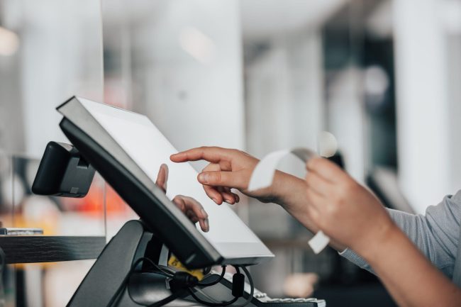 Eine Person, die einen Kassenbon in der Hand hält und ein Touchscreen-Kassenterminal in einem Geschäft verwendet, um die Einhaltung der bevorstehenden Verordnung Kassenmeldung 2025 für elektronische Kassenmeldungen zu gewährleisten.