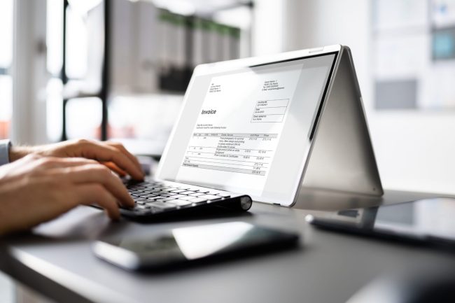 Person, die auf einer Tastatur tippt, während eine E-Rechnung (2024) auf einem Tablet im Zeltmodus auf einem Schreibtisch angezeigt wird.