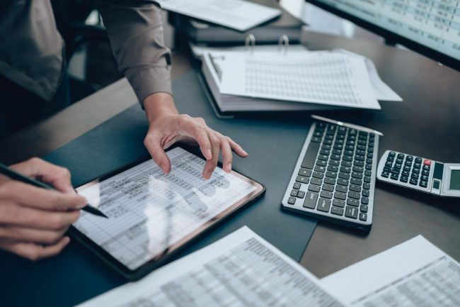 Person, die Daten auf einem Tablet überprüft und Notizen schreibt, mit einer Tastatur, Papieren, Zahlen und einem Taschenrechner auf dem Schreibtisch - Vorbereitung auf die Lohnabrechnung 2025.